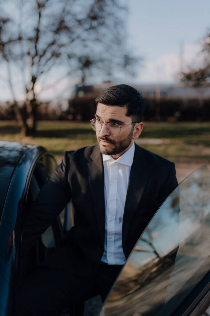 Ein Mann in einem schwarzen Anzug und mit Brille steht im Freien neben einer offenen Autotür, mit Bäumen und einem Gebäude im Hintergrund.