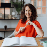 Frau mit lockigem Haar und rotem Hemd sitzt an einem Schreibtisch, lächelt, hält einen Stift und hat ein aufgeschlagenes Notizbuch vor sich. Im Hintergrund sind eine Pflanze und Schreibtischutensilien zu sehen.